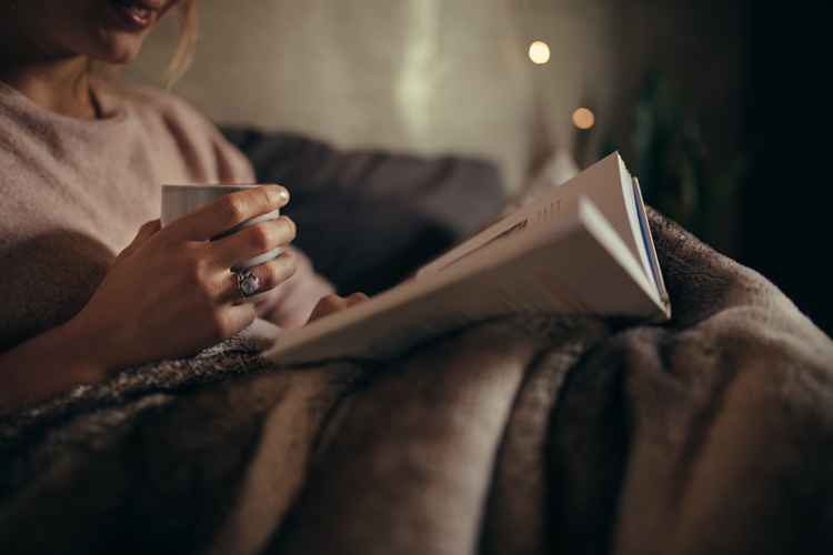 Kvinde sidder i en mørk stue med tæppe, bog og en kop te og lidt lys i baggrunden. || Hold-varmen_web.png