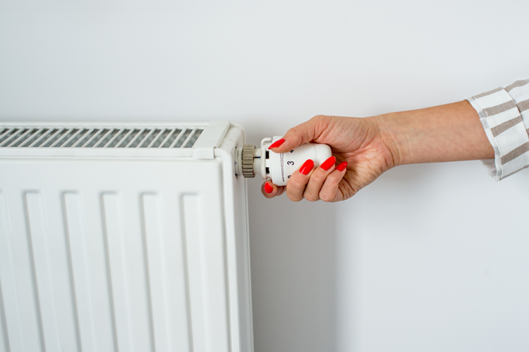 Billede af en kvindehånd med røde negle, der rækker ind i billedet fra højre og skruer på en termostat på en radiator. || Dyn Varmeregnskab Termostat Web