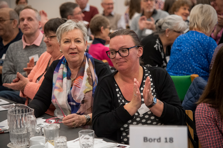 En stor gruppe af mennesker samlet i en stor sal med en kvinde i forgrunden. || Generalforsamling