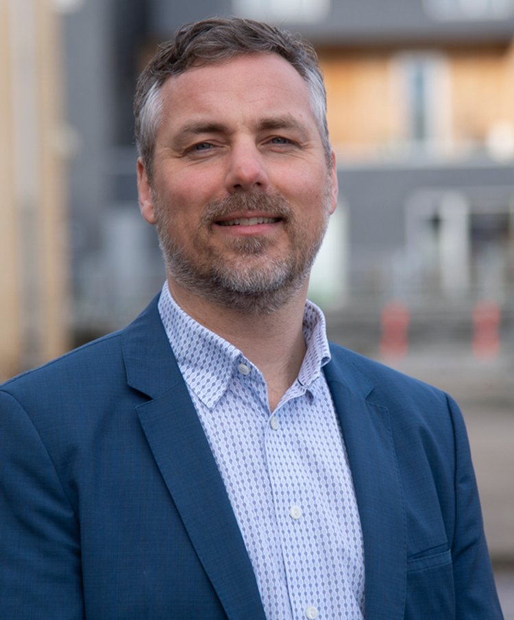 Portrætfoto af Uffe Andreasen i lyseblå skjorte og mørkeblå jakke udendørs med nogle bygninger i baggrunden. || Uffe Andreasen, Foto V Jacob Ljørring Web Lille