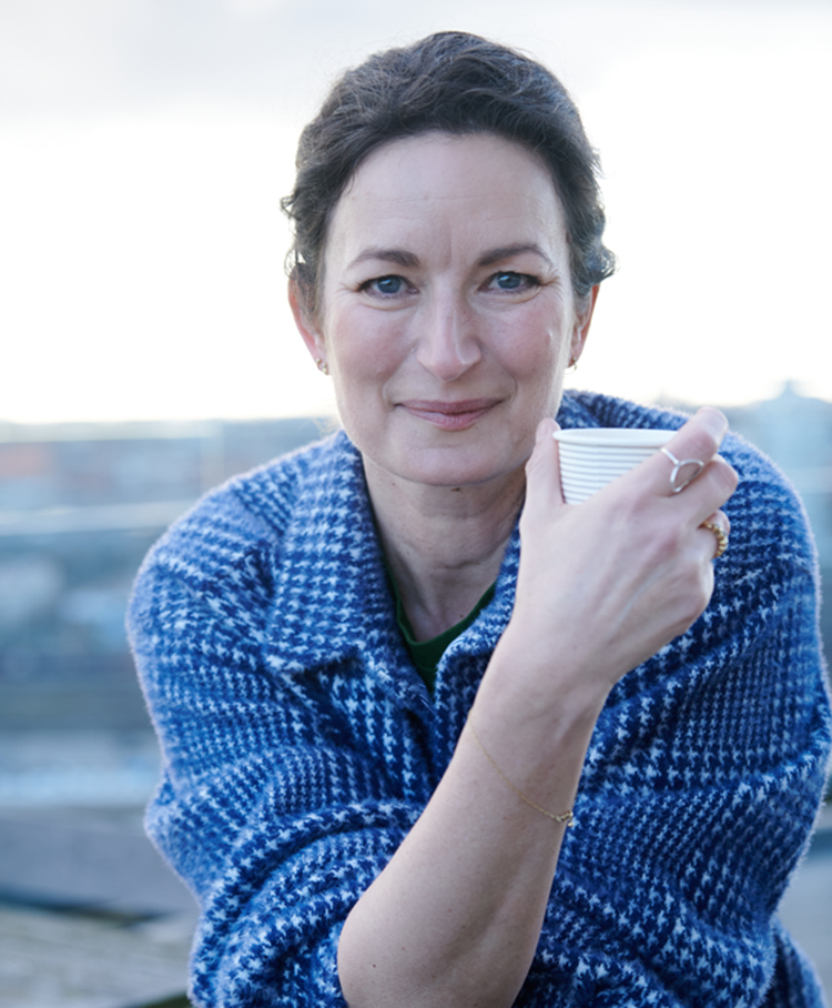 Portrætfoto af Rie Helmer siddende udenfor i en blå frakke med et krus i håneden. || Riehelmer Lille Ny