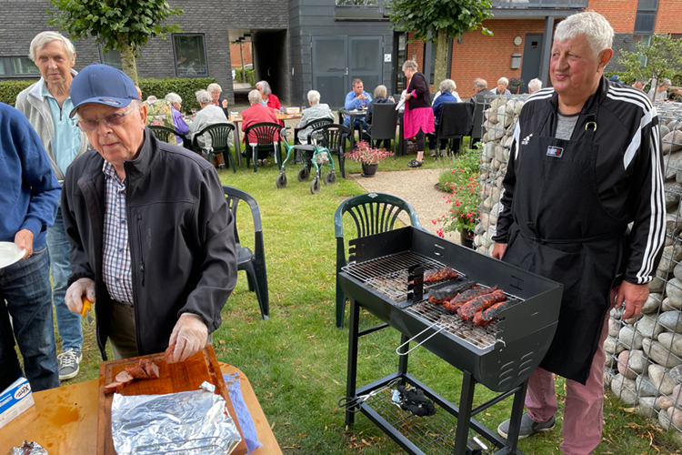 Billede af to ældre herrer, der henholdsvis passer grillen og skærer kødet udenfor. I baggrunden ses en masse mennesker sidde ved et langbord. || Hv Grill Web
