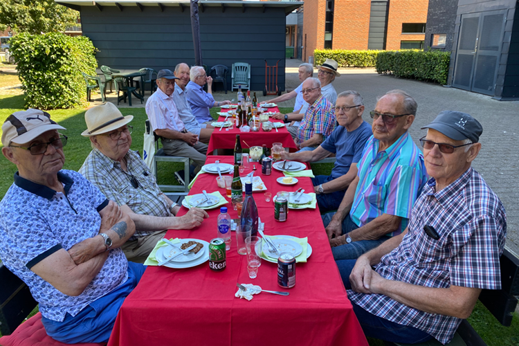 En flok ældre herrer sidder ved et langt bord med rød dug udenfor og spiser. || Hv Herresommerfest Web