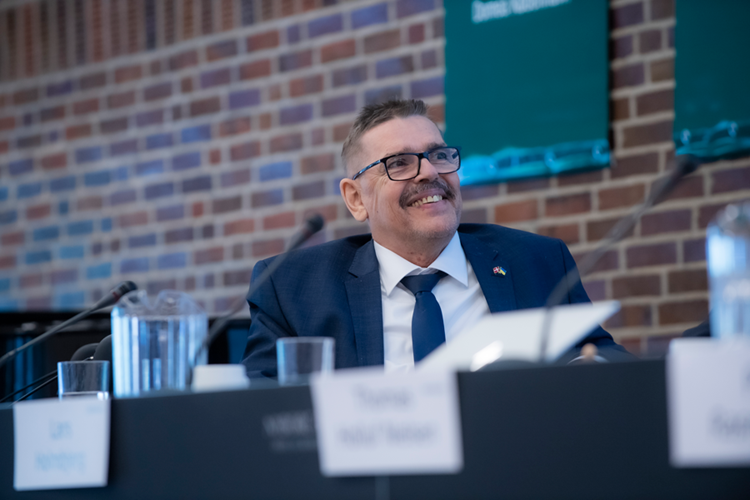 Billede af Jesper Hasemann, der sidder ved et bord på scenen. Han kigger glad op. || Jesper Hasemann GF 2022 Web