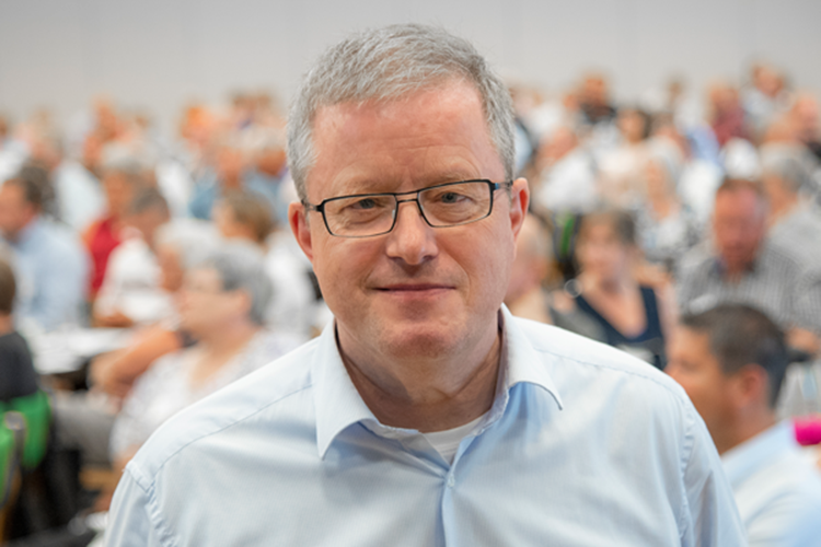 Portrætfoto af Poul Rasmussen med en siddende forsamling i baggrunden. || Poul Kundeejet Virksomhed