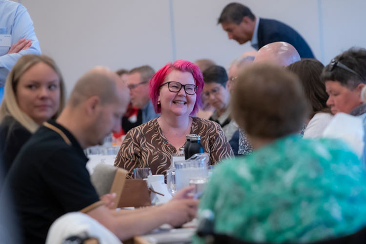 En smilende kvinde med pink hår sidder og smiler i en forsamling. || Generalforsamling Lille