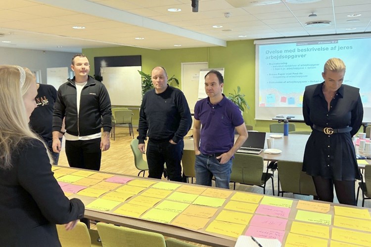 To kvinder og tre mænd står samlet om bord med en masse farvede papirer. || DDA Ovelse Arbejdsopgaver Web 900X600