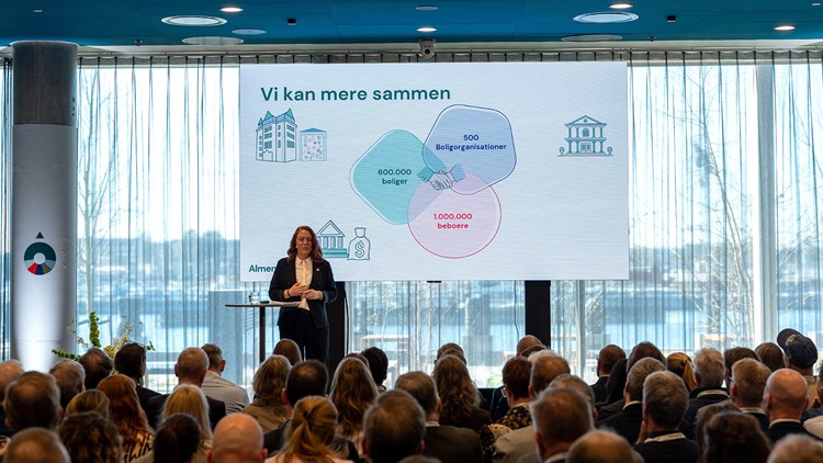 Ulla Jensen på scenen foran publikum. Bagved en stor planche, hvor der står 'Vi kan mere sammen' || Almenkompas Overdragelse 3 1600X900